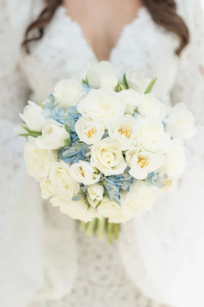 A close-up photograph of a white and blue bridal bouquet held by a bride in a white lace dress.