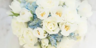 A close-up photograph of a white and blue bridal bouquet held by a bride in a white lace dress.