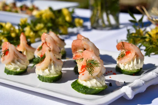 A white platter featuring shrimp appetizers served on cucumber slices with cream cheese, garnished with dill and red caviar, set on a table with yellow flowers.