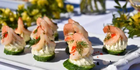A white platter featuring shrimp appetizers served on cucumber slices with cream cheese, garnished with dill and red caviar, set on a table with yellow flowers.