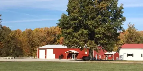 Exterior view of a red barn and white outbuildings at a country wedding venue with a large tree and grassy field.