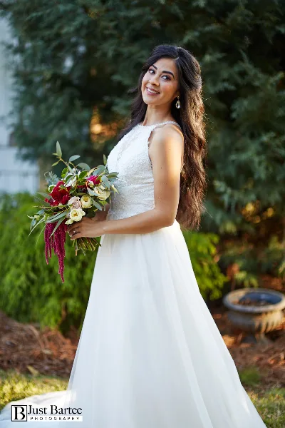 A bride in a white wedding dress holding a floral bouquet, standing outdoors with greenery in the background.