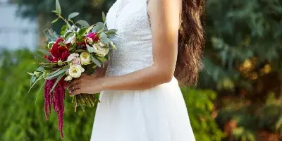 A bride in a white wedding dress holding a floral bouquet, standing outdoors with greenery in the background.