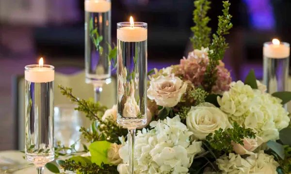 A close-up of a wedding reception floral centerpiece with white roses, hydrangeas, and tall glass pillar candles.