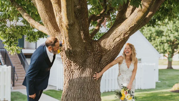 A bride and groom laughing together outdoors near a large tree, with a white building and white picket fence in the background.