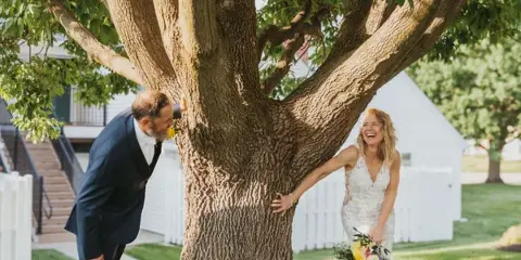 A bride and groom laughing together outdoors near a large tree, with a white building and white picket fence in the background.