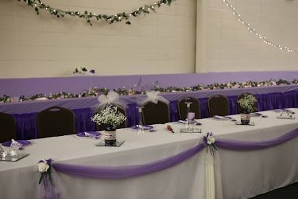 A long white table decorated with purple fabric, flowers, and bows at a wedding reception.