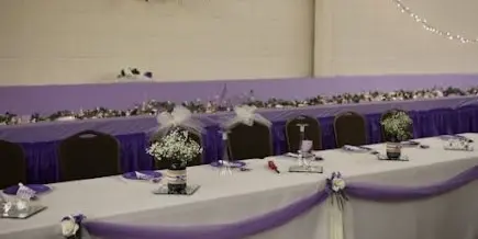 A long white table decorated with purple fabric, flowers, and bows at a wedding reception.