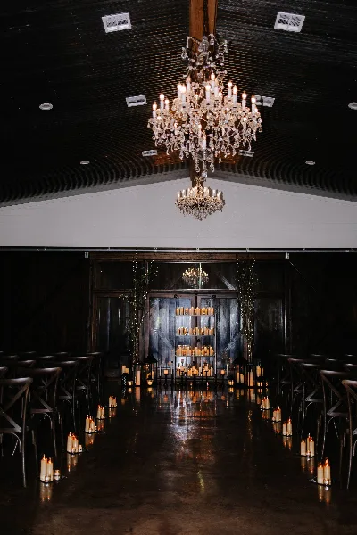 Interior view of a wedding venue featuring a long aisle lined with candles, rows of chairs, and crystal chandeliers hanging from a dark wooden ceiling.