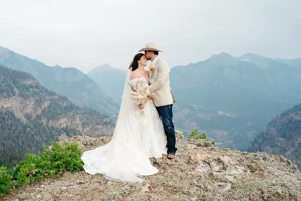 A bride and groom embracing on a rocky mountain overlook with a vast mountain range in the background.