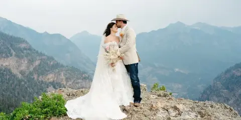 A bride and groom embracing on a rocky mountain overlook with a vast mountain range in the background.