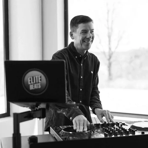 A smiling DJ in a dark shirt operating a turntable and mixer setup with an Elite Beats logo on the laptop screen.