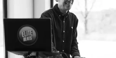 A smiling DJ in a dark shirt operating a turntable and mixer setup with an Elite Beats logo on the laptop screen.