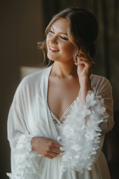 A smiling bride in a white robe with styled hair and makeup, looking down and to the side.