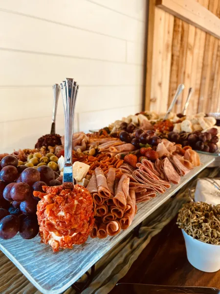 A large catering spread featuring a charcuterie board with rolled meats, cheese, grapes, and a salami ball, set on a long metal tray.