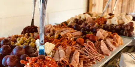 A large catering spread featuring a charcuterie board with rolled meats, cheese, grapes, and a salami ball, set on a long metal tray.