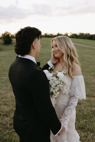 A bride and groom standing in a field, looking at each other, with the bride holding a bouquet.