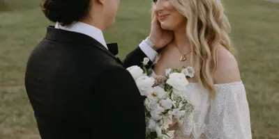 A bride and groom standing in a field, looking at each other, with the bride holding a bouquet.