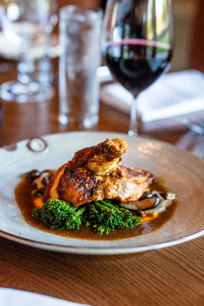 A plated dish of roasted chicken with broccolini and mushrooms served with a glass of red wine.