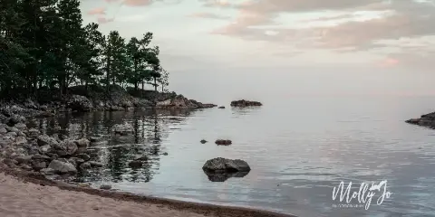 A serene Lake Superior shoreline at dusk with pine trees, rocks, and calm water, watermarked Molly Jo Photography.