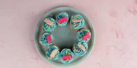 A plate of blue-frosted cupcakes topped with sprinkles and candy decorations on a pink marble surface.