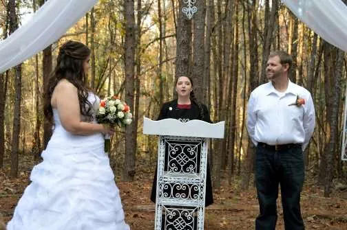 A wedding officiant stands at a podium between a bride and groom during an outdoor ceremony in a wooded area.