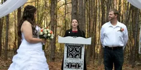 A wedding officiant stands at a podium between a bride and groom during an outdoor ceremony in a wooded area.