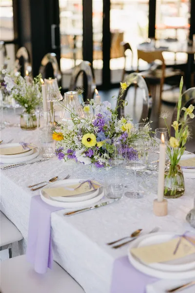 A wedding table setting featuring a long floral centerpiece with purple, yellow, and white flowers, candles, and place settings.