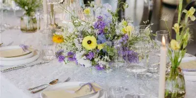 A wedding table setting featuring a long floral centerpiece with purple, yellow, and white flowers, candles, and place settings.