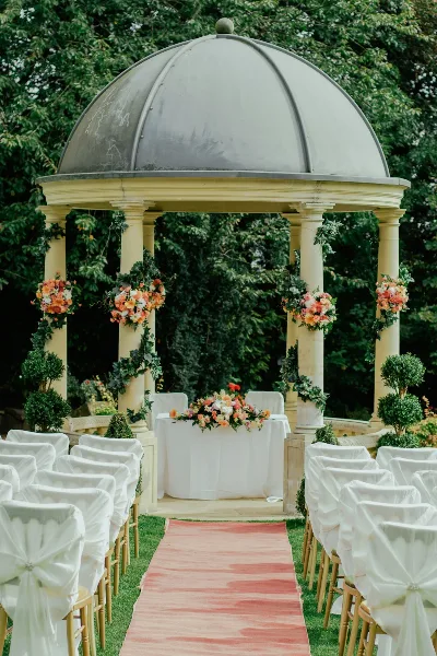 Outdoor wedding ceremony setup featuring a decorated gazebo, floral arrangements, and rows of white-covered chairs along a pink aisle.
