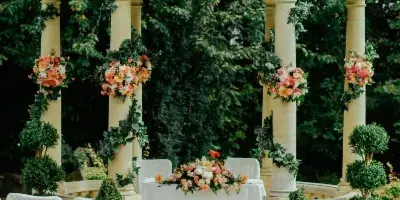 Outdoor wedding ceremony setup featuring a decorated gazebo, floral arrangements, and rows of white-covered chairs along a pink aisle.