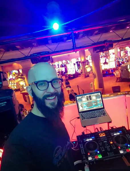 A DJ with a beard and glasses standing behind a Pioneer DJ controller and laptop at an event venue.