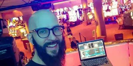 A DJ with a beard and glasses standing behind a Pioneer DJ controller and laptop at an event venue.