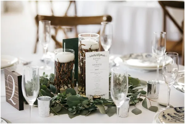 A close-up of a decorated event table featuring a greenery centerpiece, candles, glassware, and a table number sign.