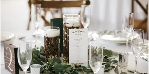 A close-up of a decorated event table featuring a greenery centerpiece, candles, glassware, and a table number sign.