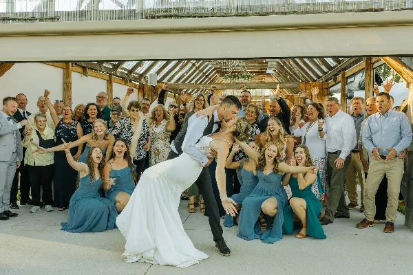 A bride and groom dip-kiss in front of a large group of cheering wedding guests inside a wooden barn structure with a glass roof.