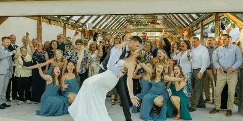 A bride and groom dip-kiss in front of a large group of cheering wedding guests inside a wooden barn structure with a glass roof.