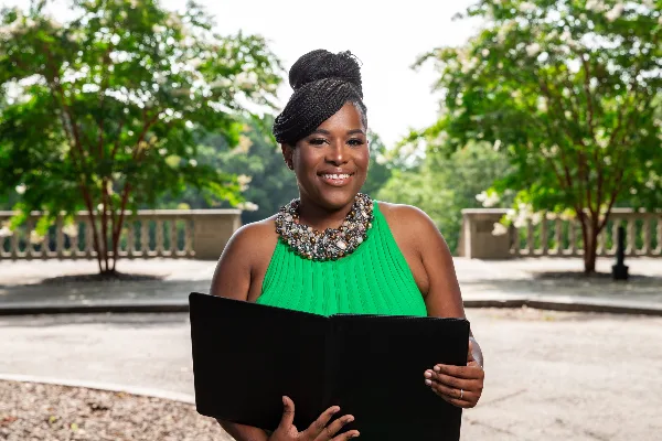A smiling woman in a green dress holding a black book outdoors