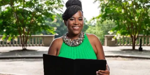 A smiling woman in a green dress holding a black book outdoors