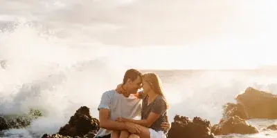 A cinematic photograph of a couple sitting on rocks by the ocean at sunset.