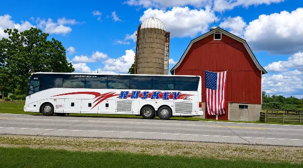 A white Huskey charter bus parked on a road in front of a red barn and silo.