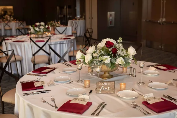 Wedding reception table set with white linens, burgundy napkins, floral centerpiece, candles, and a table number sign.