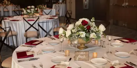 Wedding reception table set with white linens, burgundy napkins, floral centerpiece, candles, and a table number sign.