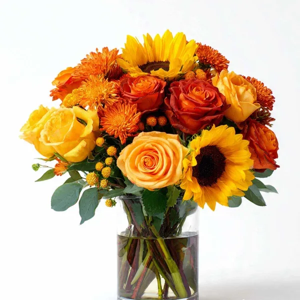 A floral arrangement of yellow and orange roses, chrysanthemums, and sunflowers in a clear glass vase.