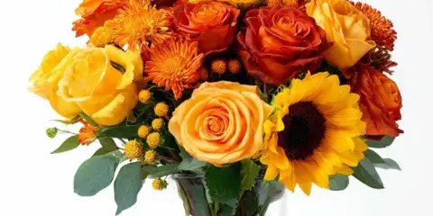 A floral arrangement of yellow and orange roses, chrysanthemums, and sunflowers in a clear glass vase.