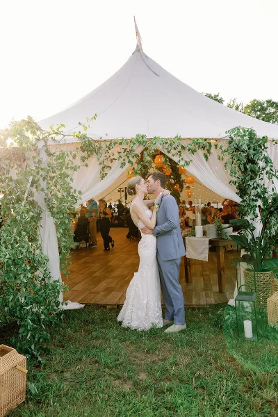 A bride and groom kissing in front of a white wedding tent decorated with greenery and lanterns.