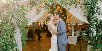 A bride and groom kissing in front of a white wedding tent decorated with greenery and lanterns.