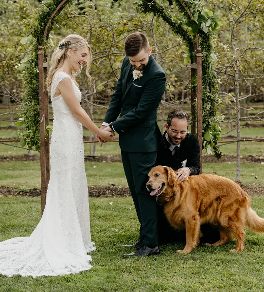 A bride and groom holding hands during an outdoor wedding ceremony with an officiant kneeling beside them and a golden retriever.