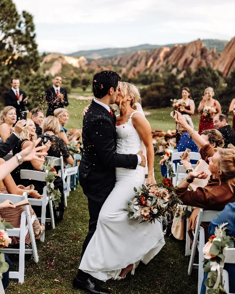 A bride and groom kiss during their wedding recessional while guests throw confetti in an outdoor venue.
