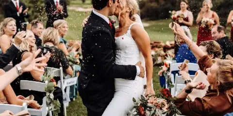 A bride and groom kiss during their wedding recessional while guests throw confetti in an outdoor venue.